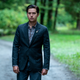 Riverdale's Jughead, dressed in a sharp suit and with slicked back hair, stands alone on a gravel road with trees in the background.